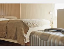 Ein schlicht eingerichtetes Schlafzimmer mit einem niedrigen Bett und einer beigefarbenen Tagesdecke vor einer holzvertäfelten Wand. Im Vordergrund ist ein silberner Koffer zu sehen.