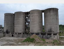 Vier große, zylindrische Beton-Silos mit sichtbarer Verwitterung und zerbrochenen Fenstern in einem verlassenen Industriegebiet, umgeben von Gras und Schutt. Ein offenes Gerüst verbindet die Silos in der Mitte, und der Himmel ist bedeckt.