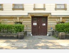 Eingangsbereich eines Gebäudes mit zwei dunkelbraunen Holztüren, verziert mit geometrischen Mustern. Umgeben von niedrigen Sträuchern und Steinbarrieren, sind an den Türen Schilder angebracht, darunter Hinweise auf einen Feuerwehrzufahrtsbereich und ein Symbol für Barrierefreiheit.