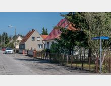 Straßenszene in einem Wohngebiet mit Einfamilienhäusern, die Giebeldächer mit roten Ziegeln haben. Links sind parkende Autos, während rechts Zäune und Vorgärten die Grundstücke einfrieden; ein Straßenschild zeigt die Kreuzung von Richard-Dehmel-Straße und Schillerstraße.