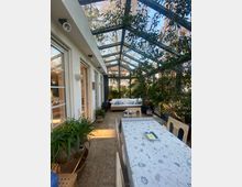 Ein lichtdurchfluteter Wintergarten mit Glasdach und Glaswänden, eingerichtet mit einem langen, gedeckten Esstisch sowie einer Sitzecke mit Sofas und Kissen. Pflanzen in Töpfen schmücken den Raum, während Bäume und blauer Himmel durch die Glaswände sichtbar sind.