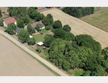 Luftaufnahme einer ländlichen Umgebung mit mehreren weiß gestrichenen Häusern, die von Bäumen umgeben sind. Neben den Gebäuden befinden sich Felder, ein Sandweg und kleinere Grünflächen.