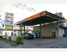 Kleine Tankstelle in einer urbanen Umgebung mit einem offenen Überdachungsbereich aus Metall. Im Vordergrund stehen Preisschilder für Kraftstoffe, im Hintergrund sind Zapfsäulen und ein kleines Gebäude mit Klinkerfassade zu sehen.