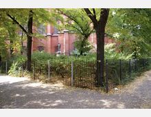 Ein mit Bäumen umgebener öffentlicher Platz mit einem schwarzen Metallzaun, dahinter dichte Vegetation und eine Statue. Im Hintergrund ist die rote Backsteinfassade eines historischen Gebäudes mit großen Fenstern zu sehen.