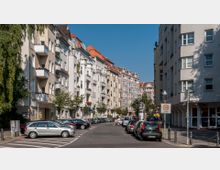 Straßenszene in einem städtischen Wohngebiet mit mehrstöckigen Altbauhäusern, die helle Fassaden und Balkone mit Blumen haben. Am Straßenrand sind geparkte Autos, während ein Schild eine Tempo-30-Zone ausweist und kleinere Bäume entlang des Gehwegs zu sehen sind.