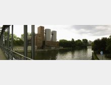 Industriegebäude aus rotem Backstein mit zwei großen, silbernen Tanks an einem Flussufer, umgeben von Bäumen. Links im Bild verläuft eine Metallbrücke, die das Flussufer verbindet.