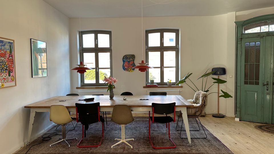 Heller Ess- oder Besprechungsraum mit großem Holztisch auf Teppich, gemischten Stühlen und zwei roten Pendelleuchten. An der Rückwand stehen zwei hohe Sprossenfenster; rechts ist eine große grüne Holztür, daneben eine Zimmerpflanze und ein Sessel.