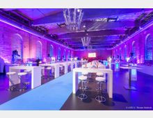 Großer Veranstaltungsraum mit Ziegelwänden, farbiger Beleuchtung in Blau und Lila, hohen weißen Stehtischen mit passenden Hockern und einer zentralen blauen Bodenbahn. Die Decke zeigt sichtbare Betonträger, und moderne Kronleuchter hängen von oben.