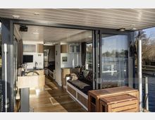Innenraum eines modernen Hausboots mit Holzlaminatboden, Sitzecke, kleiner Küche und Steuerstand. Große Fensterfronten und eine Terrasse bieten Blick auf das umliegende Gewässer und die Uferlandschaft.