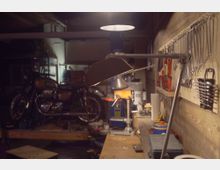 Innenansicht einer Werkstatt oder Garage mit Werkbank im Vordergrund und einer Werkzeugwand mit aufgehängten Schraubenschlüsseln und Zangen. Im Hintergrund stehen ein Motorrad und eine Ständerbohrmaschine unter gedämpfter Deckenbeleuchtung.