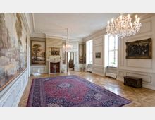 Ein prunkvoller historischer Raum mit aufwendig verziertem Parkettboden, einem großen roten Teppich, zwei Kronleuchtern und hohen Fenstern. Die Wände sind mit Gemälden und Holzvertäfelungen dekoriert, ein Kamin mit vergoldetem Rahmen steht zentral an der Wand.