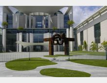 Moderne Regierungsgebäude mit symmetrischem Aufbau, Glasfassaden und Säulen; im Vordergrund steht eine große abstrakte Skulptur aus Metall, umgeben von gepflegten Rasenflächen und einem Zaun.