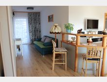 Wohnzimmer mit Holzboden, einem blauen Sofa, dekorativen Vorhängen und einer hohen Holztheke mit Stühlen. Im Hintergrund befinden sich ein Fenster mit durchscheinenden Vorhängen sowie ein Fernseher an der Wand.