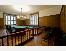 Raumansicht eines Sitzungssaals mit holzvertäfelten Wänden, einem ovalen Konferenztisch mit grüner Tischdecke, umgeben von Stühlen. Der Raum hat große Fenster, einen Parkettboden und eine zentrale Deckenleuchte mit mehreren Leuchtarmen.