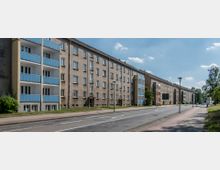 Straßenszene mit einer Reihe von mehrstöckigen Wohnhäusern im Plattenbaustil, teils mit blauen Balkonen. Im Vordergrund verläuft eine zweispurige Straße mit Gehwegen und vereinzelt stehenden Straßenlaternen, flankiert von Bäumen.