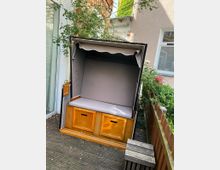 Ein Strandkorb mit grauem Stoffdach und Sitzpolster steht auf einer Holzterrasse, umgeben von Pflanzen. Im Hintergrund sind Gebäudefassaden, ein Fenster und ein Zaun sichtbar.