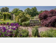 Ein gepflegter Garten mit einer Holzbank auf einem gepflasterten Weg, umgeben von bunten Blumen, Sträuchern und einem roten Ahornbaum; im Hintergrund ein von Pflanzen bewachsener Pavillon und hohe Bäume.