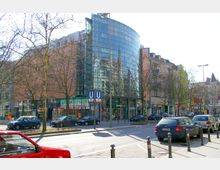 Straßenszene in einer Stadt mit einem modernen Gebäude mit einer großen, gebogenen Glasfassade, die umliegende Gebäude reflektiert. Im Vordergrund parken Autos entlang der Straße, und ein U-Bahn-Schild für die Linien U2 und U3 ist sichtbar.