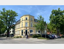 Gelbes, zweistöckiges Eckhaus im klassischen Stil mit weißen Fensterrahmen und einem Schieferdach, umgeben von Bäumen. Vor dem Gebäude befinden sich Sonnenschirme, darunter Tische, sowie ein kleiner Parkplatz mit geparkten Autos.