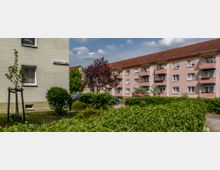 Mehrfamilienhäuser einer Wohnsiedlung mit hellen Fassaden und roten Dächern, umgeben von gepflegten Büschen und kleinen Bäumen. Im Vordergrund eine Rasenfläche und ein Straßenschild mit der Aufschrift "Wagnerstraße 1a-1c", im Hintergrund geparkte Autos.