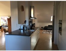 Wohnraum mit offener Gestaltung, bestehend aus einer modernen Küche mit weißer Einbauküche und einer schwarzen Arbeitsplatte im Vordergrund. Im Hintergrund sind ein Wohnzimmer mit einer Couch, Teppich und Couchtisch sowie ein Essbereich mit Tisch und Stühlen zu sehen, erhellt durch Dachfenster und eine Glastür, die auf eine Terrasse führt.