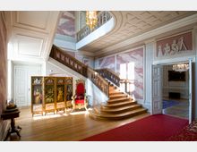 Innenraum eines historischen Gebäudes mit hölzerner, geschwungener Treppe und verzierten Geländern, dekorativen Wandmalereien und Stuckarbeiten. Im Vordergrund ein roter Thronsessel sowie ein dekorativer Paravent mit Porträts, auf dem Boden ein roter Teppich.
