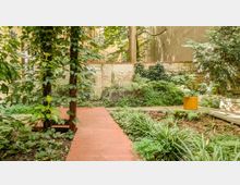 Ein kleiner, bewaldeter Garten mit Holzstegen, dichtem Pflanzenbewuchs und einer Ziegelmauer im Hintergrund. Im Vordergrund stehen Bäume und Sträucher, seitlich befindet sich ein leuchtend orangefarbener Pflanzentopf.