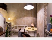 Ein kleiner, gemütlicher Raum mit grob verputzten Wänden, einem runden Tisch, zwei Stühlen und einer weißen Bank. Auf dem Tisch stehen Keramikdekorationen und eine Kerze, daneben befindet sich ein Regal mit Haushaltsgegenständen, während eine Hängelampe den Raum beleuchtet.