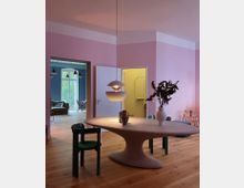 Moderner Essbereich mit hellrosa Wänden, einem ovalen Tisch mit organischer Form und grünen Stühlen auf einem Holzfußboden. Im Hintergrund sind angrenzende Räume mit blauen Wänden, großen Fenstern und einer gelben Tür zu sehen. Eine Hängelampe und dekorative Gegenstände auf dem Tisch ergänzen den Raum.