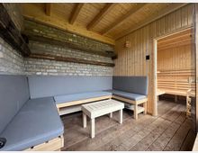 Ein gemütlicher Ruhebereich mit Holzboden und gemauerten Wänden, ausgestattet mit einer Eck-Sitzbank mit grauen Polstern und einem kleinen Holztisch; rechts ist ein Durchgang zu einer Sauna mit Holzbänken sichtbar.