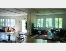 Großer, heller Raum mit Holzboden, großen Fenstern und Blick auf grüne Pflanzen draußen. Im Raum stehen ein Klavier, ein Schlagzeug, Gitarren, ein Sofa, ein Fahrrad und ein Schaukelpferd, mit weiteren künstlerischen und musikalischen Elementen verteilt.