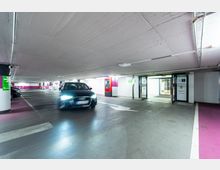 Ein beleuchtetes Parkhaus mit niedriger Decke, einem fahrenden Auto im Vordergrund und klar markierten Fahrspuren. Im Hintergrund sind ein Ticketautomat, Schilder für den Ausgang und Glastüren zu sehen.