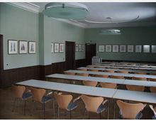 Ein Seminarraum mit langen weißen Tischen und holzfarbenen Stühlen in mehreren Reihen. Die Wände sind hellgrün gestrichen und mit gerahmten Zeichnungen dekoriert, während die dunkle Holzvertäfelung den unteren Wandbereich einnimmt; zwei große, runde Deckenleuchten und ein Rednerpult sind ebenfalls sichtbar.