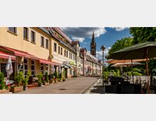 Eine gepflasterte Fußgängerzone mit Sitzbereichen unter Sonnenschirmen, flankiert von zweigeschossigen Gebäuden mit Restaurants und Geschäften, deren farbige Fassaden und Markisen auffallen. Im Hintergrund ragt ein Kirchturm in den blauen Himmel mit Wolken, während Bäume auf der rechten Seite die Szene einrahmen.