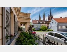 Blick auf einen kleinen Platz mit gepflasterter Straße, umgeben von Wohnhäusern mit roten und hellen Dächern. Im Hintergrund ragen die Türme einer gotischen Kirche in den Himmel, im Vordergrund sind ein Zaun, Pflanzen und ein geparktes Auto zu sehen.