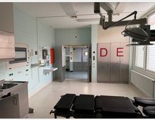 Ein steriler Raum in einem Krankenhaus oder einer medizinischen Einrichtung mit hellgrünen gefliesten Wänden, einer Waschstation, einem Edelstahlspülbecken, medizinischen Wandanschlüssen und einem Operationsstuhl. Im Hintergrund sind Edelstahl-Doppeltüren mit roten Buchstaben "DE" sowie ein Verbindungsgang sichtbar.