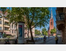 Straßenszene mit Blick auf eine rote Backsteinkirche mit hohem spitzen Turm im Hintergrund, umgeben von Bäumen und klassischen Wohngebäuden. Im Vordergrund sind ein Werbesäule, Gehwege, parkende Fahrräder, Fußgänger und ein Radfahrer zu sehen.