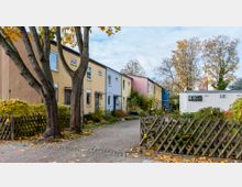 Reihenhäuser mit bunten Fassaden in einer Wohnsiedlung, umgeben von Gartenanlagen und Bäumen mit Herbstlaub. Ein gepflasterter Gehweg und ein Holzzaun säumen den Vordergrund.