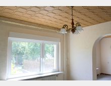 Innenraum eines Hauses mit einer großen Fensterfront, die auf einen Garten mit Bäumen und einem Holzzaun blickt. Die Decke ist holzvertäfelt, und ein antiker Kronleuchter hängt von der Decke, während ein Heizkörper unter dem Fenster angebracht ist. Rechts führt ein bogenförmiger Durchgang in einen weiteren Raum.