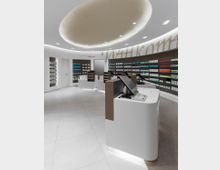 Moderne Apothekeneinrichtung mit weißen Wänden, einem geschwungenen Deckenelement mit integriertem Licht, und Regalen voller farbiger Arzneimittelverpackungen. Im Vordergrund befinden sich abgerundete Kassenarbeitsplätze mit Bildschirmen und Tastaturen.