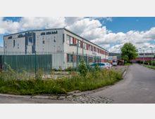 Industriegebäude mit abblätternder Fassade und roten Fensterrahmen, umgeben von einem grünen Metallzaun und wild wachsender Vegetation. Im Vordergrund verläuft eine asphaltierte Straße mit vereinzelten geparkten Autos, im Hintergrund weitere Gebäude und ein Baum.