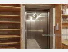 Ein moderner Aufzug mit Edelstahlwänden in einem Raum mit Sichtbeton und offenen Holzregalen auf beiden Seiten. Rechts neben dem Aufzug befindet sich eine Gegensprechanlage an der Wand.