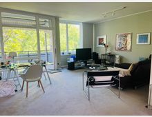 Helles, modern eingerichtetes Wohn- und Esszimmer mit großen Fensterfronten und offener Balkontür zum Balkon mit Blick auf grüne Bäume. Im Raum stehen ein Glastisch mit Stühlen, eine Sitzecke mit Sofa und Sessel sowie ein Fernseher; an der Wand hängen gerahmte Kunstwerke.