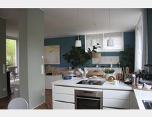 Moderne Wohnküche mit weißer Einbauküche, integriertem Herd und Hängelampen. Im Hintergrund ist ein gemütlicher Wohnbereich mit blauer Wand, Pflanzen, dekorativen Elementen und großen Fenstern zu sehen.