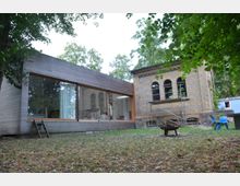 Modernes Gebäude mit Holzverkleidung und großen Glasfenstern, neben einem historischen Backsteingebäude mit Rundbogenfenstern. Im Vordergrund befindet sich eine Wiese mit Laub, einem Metall-Feuerschalenständer und Kinderspielstühlen.