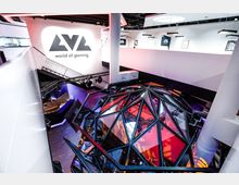 Moderner Gaming-Bereich mit futuristischem Design, einschließlich einer kuppelförmigen Struktur aus schwarzen Metallstreben und Glas, beleuchtet in bunten Farben. Im Hintergrund ist ein großes Schild mit der Aufschrift "LVL world of gaming" an einer weißen Wand zu sehen.