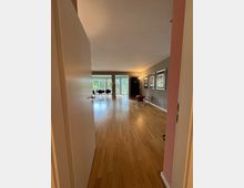 Blick in einen offenen Wohnraum mit Holzboden, großen Fenstern im Hintergrund, die Blick auf einen Garten bieten, und einer seitlich dekorierten Wand mit gerahmten Bildern. Im Raum befinden sich moderne Möbel wie Stühle und Tische.