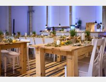 Ein modern eingerichteter Veranstaltungsraum mit Holztischen, weißen Stühlen und dekorativen Elementen wie Zitronen, Kerzen und grünem Laub auf den Tischen. Der Boden besteht aus gestreiftem Holz, während hängende Lampen und eine Bar im Hintergrund die Atmosphäre ergänzen.