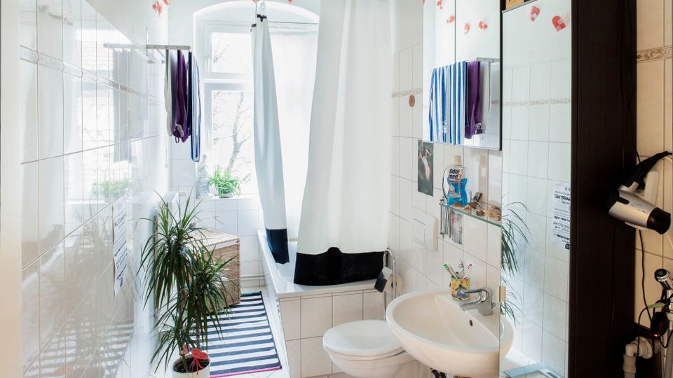 Bright, narrow tiled bathroom with a bathtub and white shower curtain at the far end beneath a large window. A wall-mounted sink with mirrored cabinet, towels, a striped floor rug, and potted plants line the space.