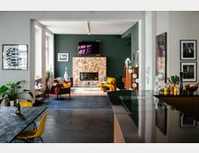 Offener Wohnraum mit modernem und farbenfrohem Interieur, bestehend aus einem Essbereich mit einem dunklen Holztisch und gelben Stühlen sowie einem Wohnzimmer mit grün gestrichener Wand, Kamin, Leder- und Stoffsesseln und bunten Dekorelementen. Im Vordergrund ist eine schwarze Küchenarbeitsplatte sichtbar, und der Raum ist durch große Fenster gut beleuchtet.